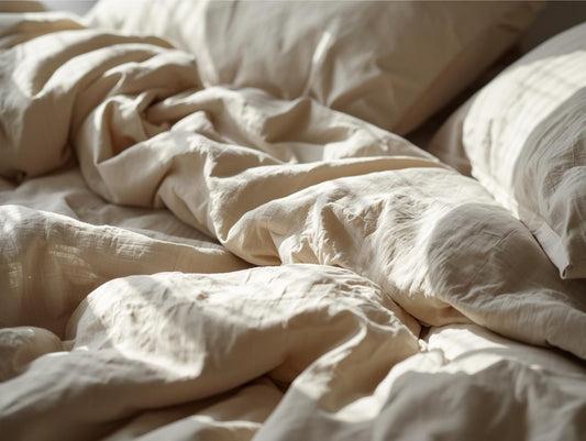 Nahaufnahme eines ungemachten Bettes mit hochwertiger beiger Leinenbettwäsche im sanften Morgenlicht, das die Textur des Stoffes hervorhebt.